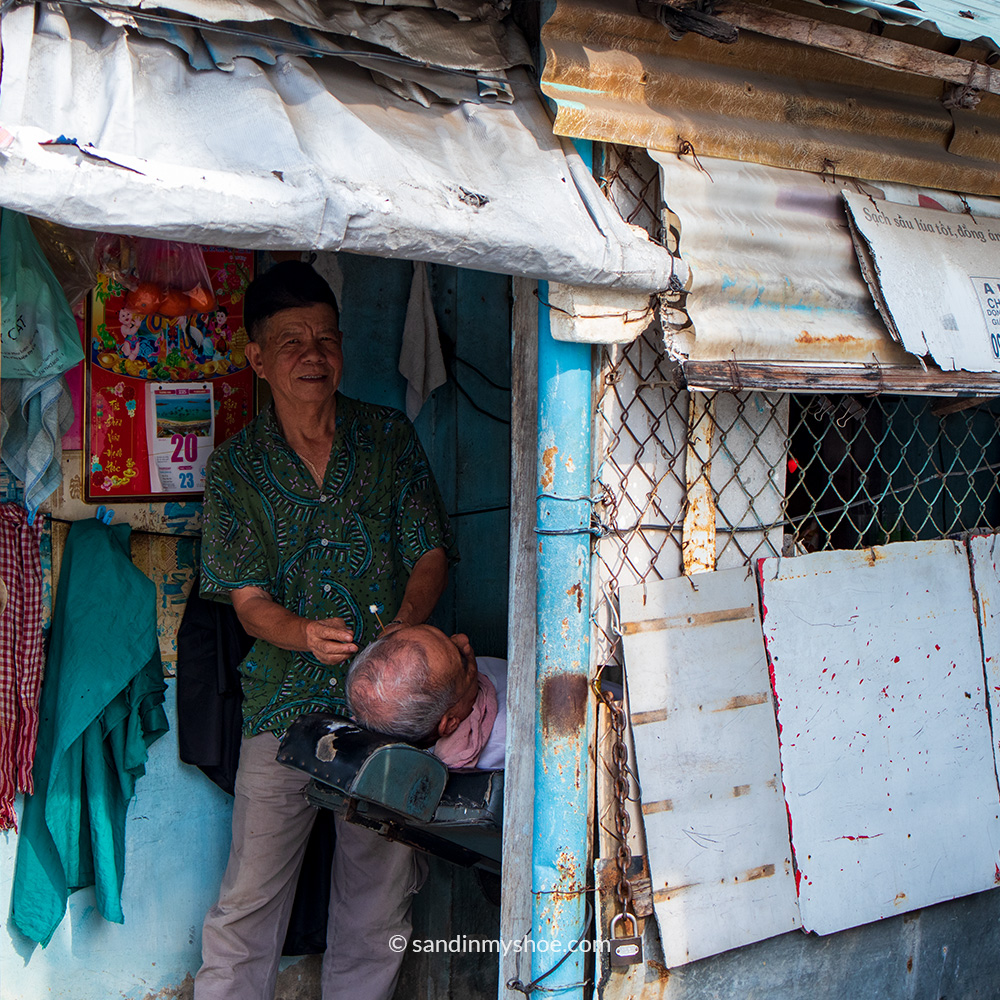 A small barbershop with a reclining chair, a plastic sheet used as a door, and a window covered by a standard metal fence.