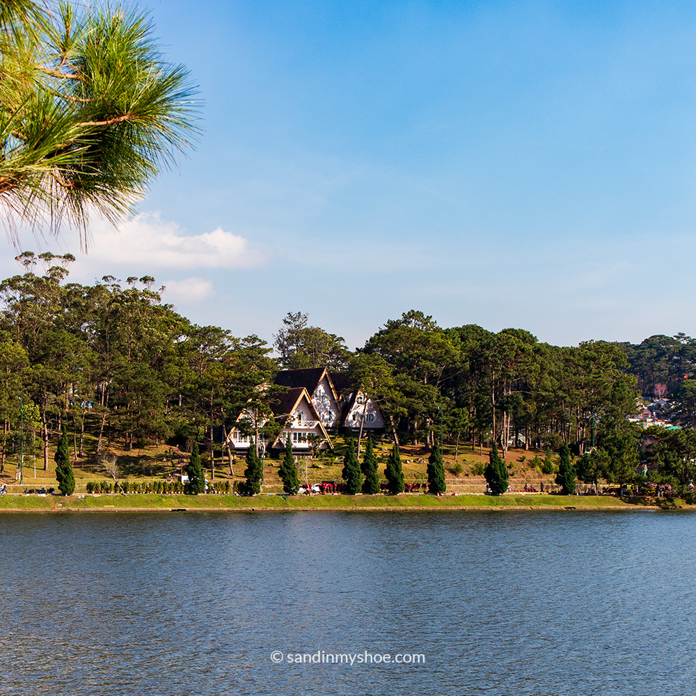 A view of Xuan Huong Lake with calm water and cabin‑style houses lining the shore.