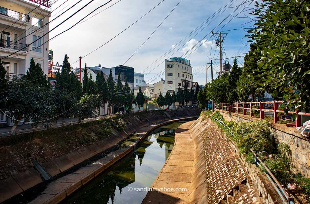 The canal in Dalat on a bright sunny day, with reflections on the water and greenery along the banks.