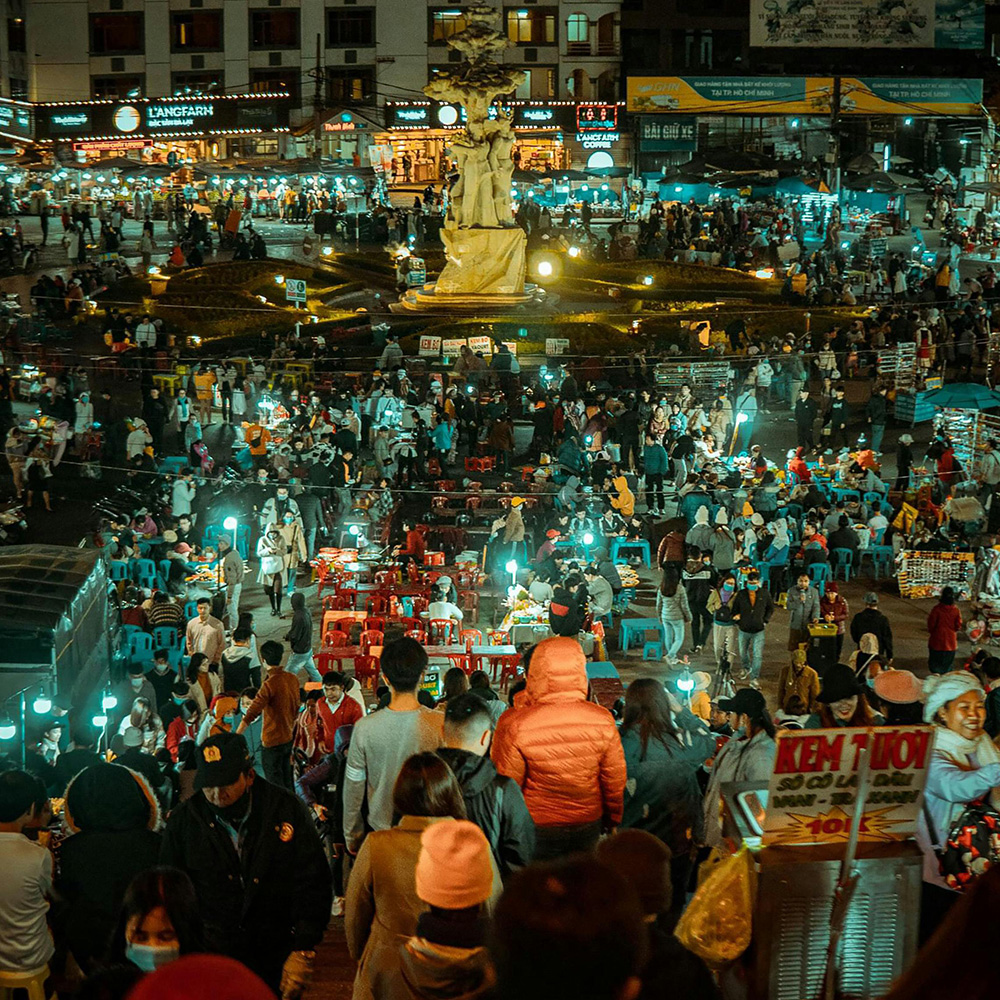 Night market scene in Dalat with food stalls, bright lights, and crowds walking through the central square.
