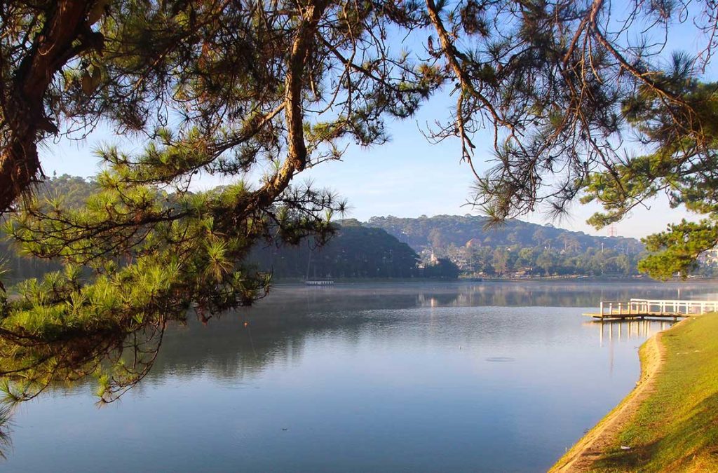 View of Xuan Huong Lake in Da Lat with calm water, pine trees along the shore, and a small wooden pier extending into the lake.