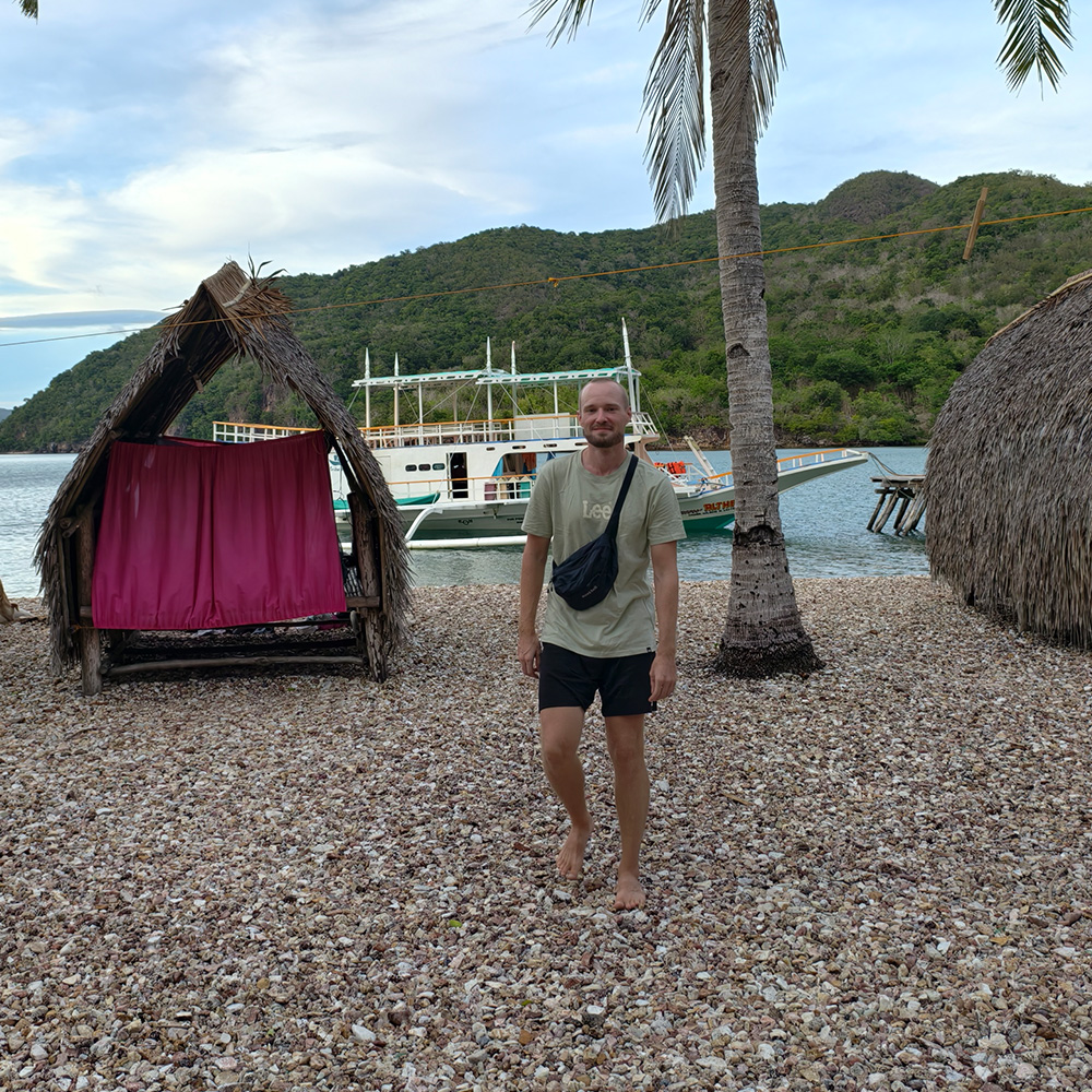 Petteri standing next to a simple bamboo hut next to the sea with the boat in the background.