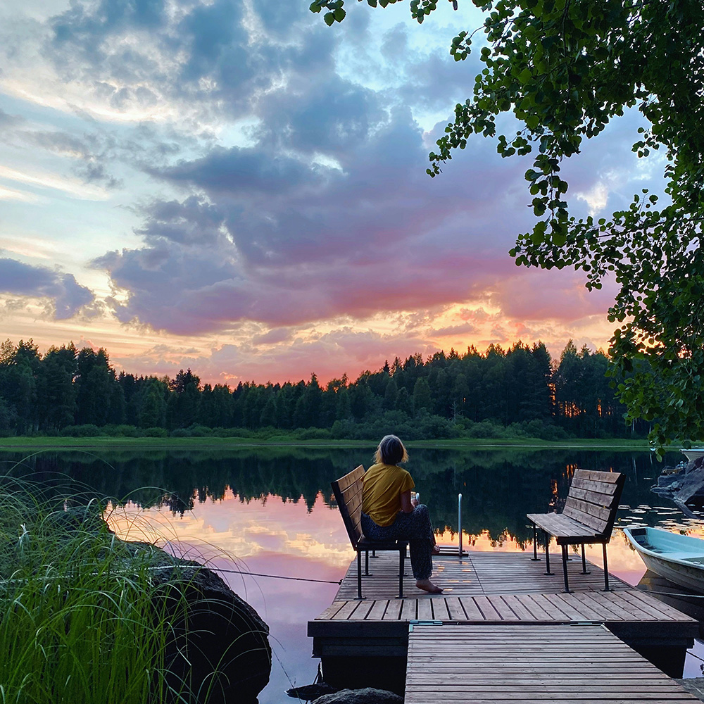 Woman looking across a calm lake on a late summer evening, surrounded by forest and soft light.